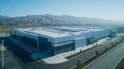 The large semiconductor fabrication plant features sleek architecture, surrounded by mountains, with clear skies overhead. It sits in a spacious environment with ample parking available