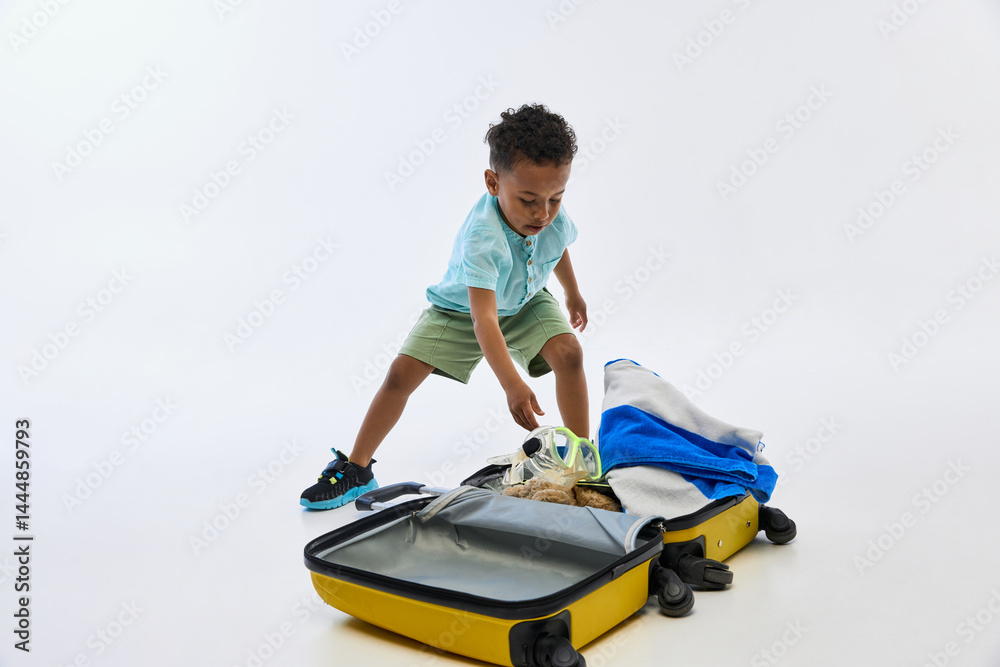 Playful and curious little African-American boy packs snorkeling gear and towels into yellow suitcase while preparing for travel on white background. Concept of childhood, adventure, vacation, play. Playful and curious little African-American boy packs snorkeling gear and towels into yellow suitcase while preparing for travel on white background. Concept of childhood, adventure, vacation, play.