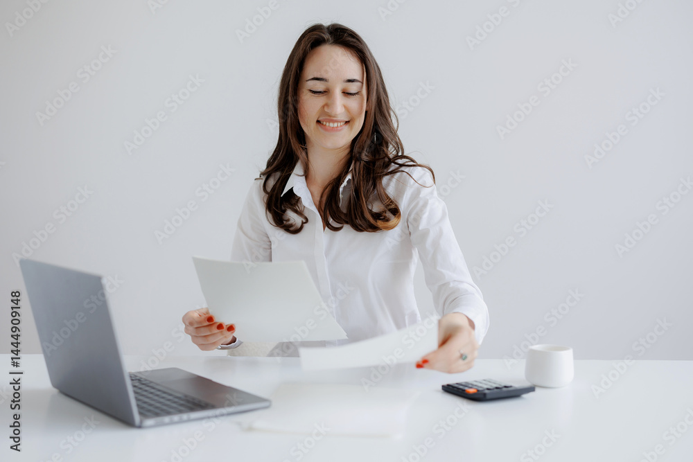 Young woman reviewing documents at a white desk with a laptop and calculator during a daytime office setting