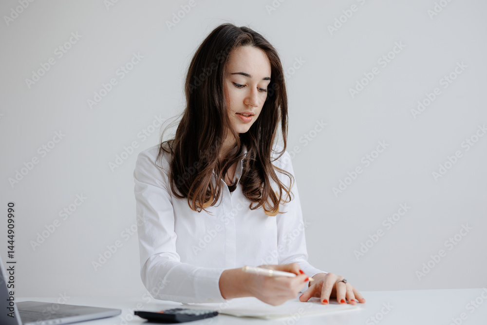 Young woman writing notes at a minimalist workspace with a laptop and calculator during a calm afternoon