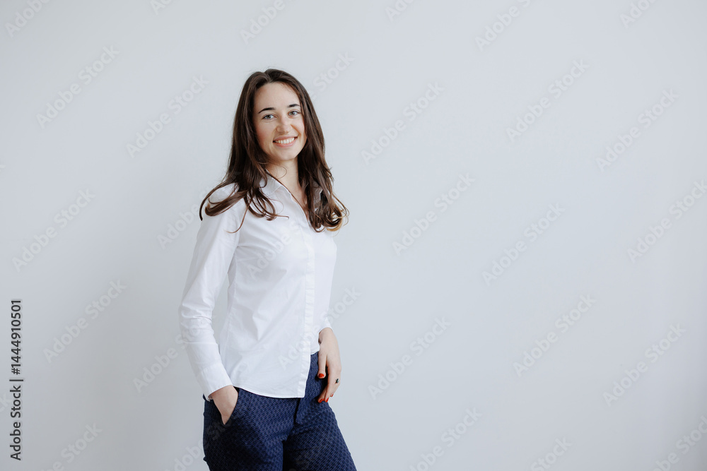 Young woman in a white shirt and navy pants smiling while standing against a light gray wall in a bright indoor space