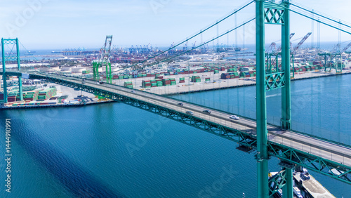 High-angle view of the iconic Vincent Thomas Bridge spanning the Port of Long Beach in California. The turquoise suspension bridge crosses over calm blue water, with stacks of shipping container