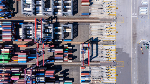 A straight-down aerial photo of shipping cranes operating over a dock filled with colorful cargo containers. The image highlights the symmetry and industrial activity of a modern shipping port.