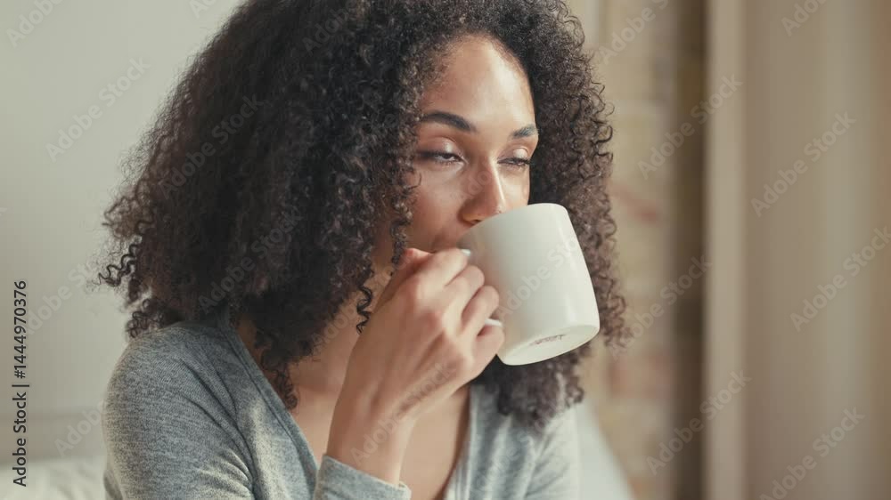 Radiant African American woman smiling warmly while holding mug in hands. Taking sip of drink. Appearing cheerful and refreshed while enjoying pleasant morning in peaceful bedroom setting.