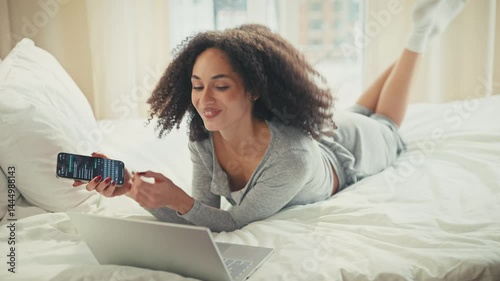Wallpaper Mural ocused African American woman finishing typing on laptop then grabbing phone with one hand. Lying on stomach and replying to message while multitasking between devices during relaxed morning in bed. Torontodigital.ca