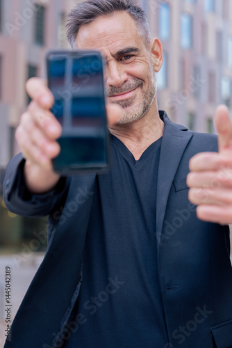 Smiling man holding smartphone towards the camera and giving a thumbs up outdoors. He wears a black jacket and stands in a modern urban space. Concept of approval, technology, and confidence. Vertical