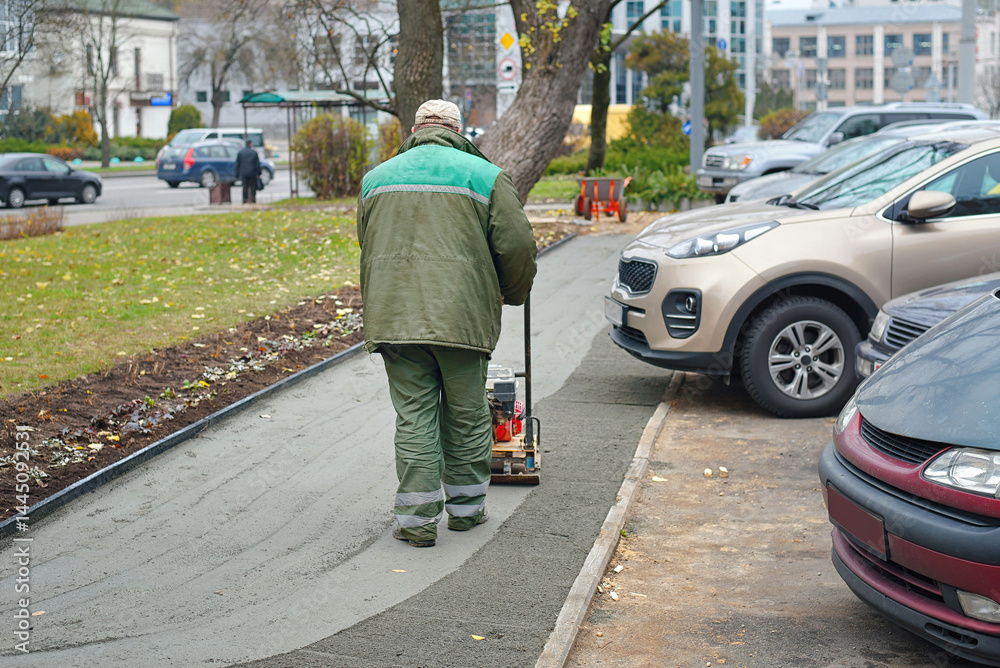 Construction worker operating compactor on fresh concrete sidewalk beside parked cars, urban roadwork maintenance to level and finish pedestrian pavement. Worker compressing fresh cement sidewalk