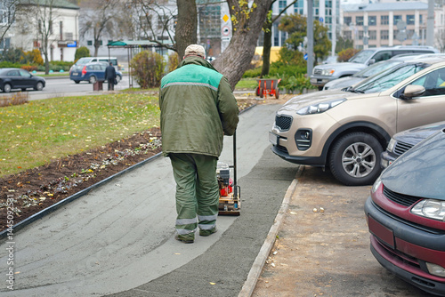 Obraz na płótnie Construction worker operating compactor on fresh concrete sidewalk beside parked cars, urban roadwork maintenance to level and finish pedestrian pavement. Worker compressing fresh cement sidewalk w salonie