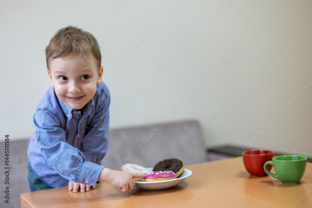 Cheerful boy reaching for donuts on table. Smiling young boy in blue shirt playfully reaching for a plate of colorful donuts on a wooden table with cups nearby. Cheerful boy reaching for donuts on table. Smiling young boy in blue shirt playfully reaching for a plate of colorful donuts on a wooden table with cups nearby.