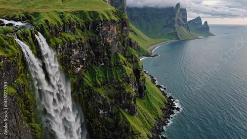 Waterfall plunging from grass-covered cliff into turquoise sea with distant peaks
