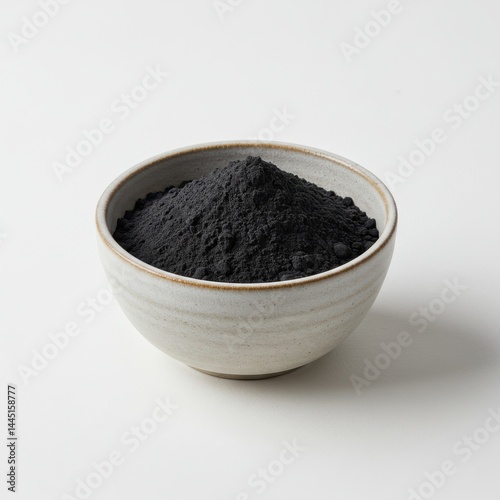A ceramic bowl filled with a heap of dark gray powder on a plain white surface in a studio setting