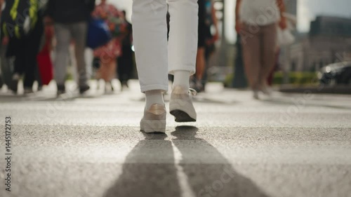 Back view following shot female feet steps walking pedestrian crossing street, sunny day, young woman wearing white pants and sneakers goes crosswalk. Low angle people legs stepping zebra asphalt