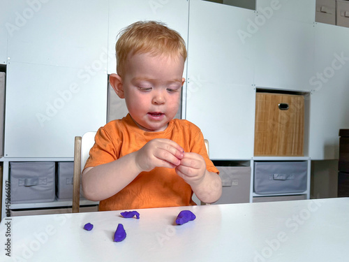 A little red-haired boy is sitting and sculpting from colored plasticine at a table in a room. Developing fine motor skills at the table
