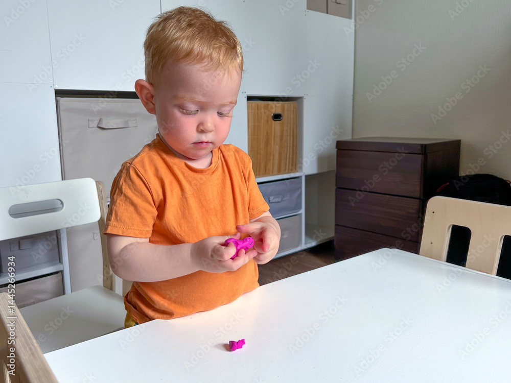 A red-haired boy in an orange T-shirt stands in front of a table and sculpts from plasticine. Early development of a child at home