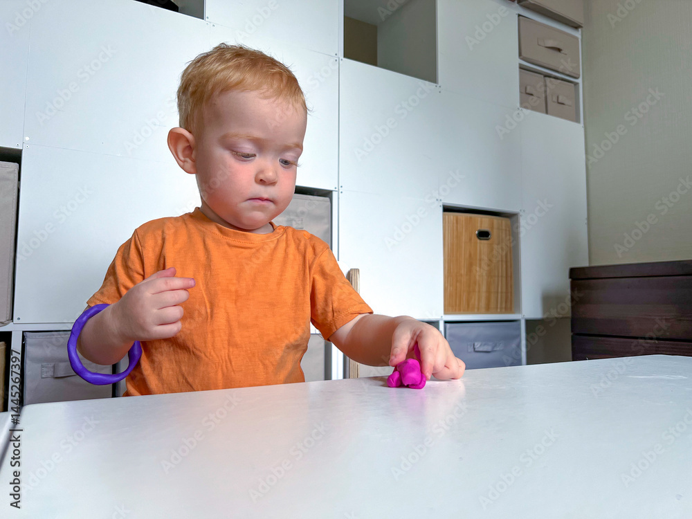 A little red-haired boy with a ring made of plasticine on his hand is playing at a table with plasticine. Early development of fine motor skills, creativity