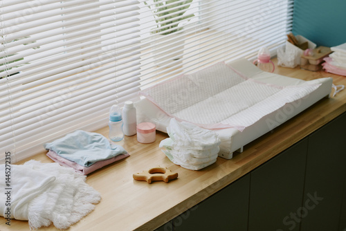 Clean and organized baby changing station with diapers, wipes, clothes, and changing pad arranged on wooden countertop next to window with blinds partially open