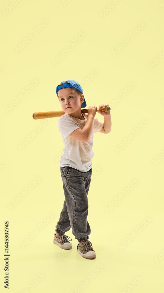 Fototapeta premium Young boy in dressed casually with blue cap preparing to hit baseball with bat against pastel yellow background. Concept of sport, hobby, childhood, wellness, vacation, energy, happiness, growth.