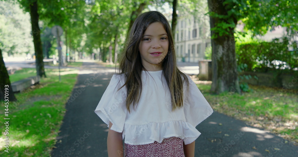 Naklejka premium Happy girl standing on a sunny tree-lined path in a green park, smiling warmly at the camera, wearing a white blouse and patterned skirt, enjoying a beautiful day