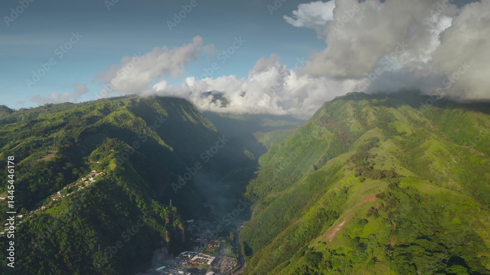 Obraz premium Stunning aerial view of lush green valley with small village nestled amidst towering mountains, with clouds over hill peaks, creating a mesmerizing landscape in the heart of Tahiti, French Polynesia