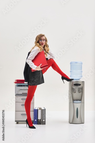 Quadro em tela Woman with red folder striking exaggerated pose with leg on water cooler beside printer in surreal corporate setting against white background
