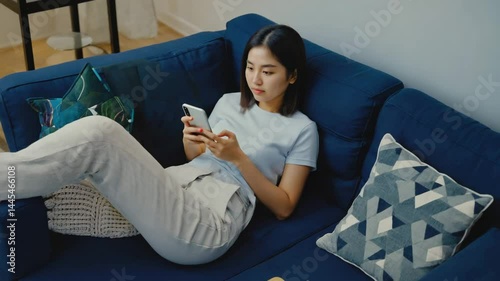 Smiling young woman sitting on a sofa at home uses her mobile phone with a smile, enjoying leisure time with technology
