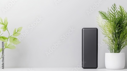 Black electronic device stands on a white table next to a potted green plant against a minimalist grey wall.