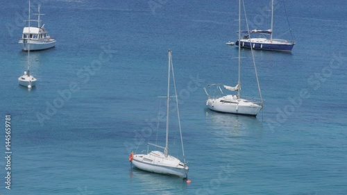 Several sailboats anchored in calm blue sea with mountains and coastal town in background. Sunny summer day in Mallorca, peaceful maritime scene, themes of travel, vacation, sailing, and nature.