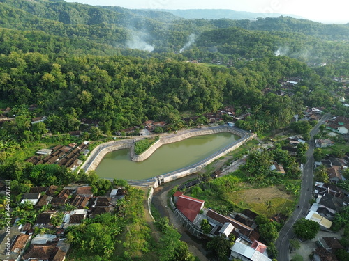 Special Region of Yogyakarta, Indonesia - April 25, 2025 : Aerial View of a Rural Reservoir Surrounding Lush Green Forest and Houses photo