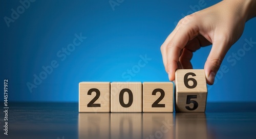 Hand placing a wooden block with number 6 to change year 2025 to 2026 on a blue background.