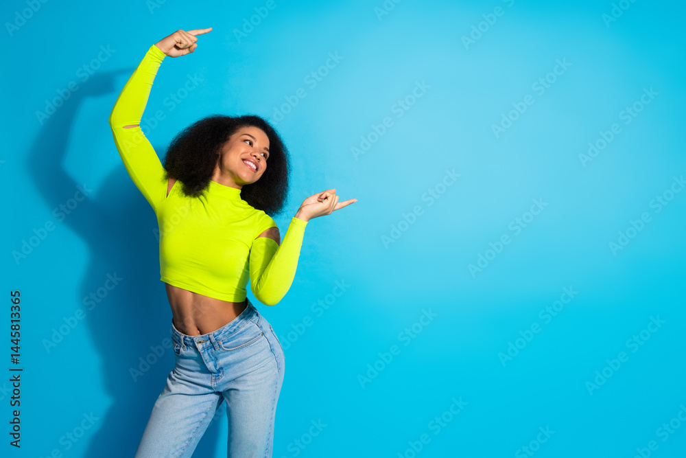 Joyful young woman wearing neon green top, expressing happiness with finger gestures against blue background