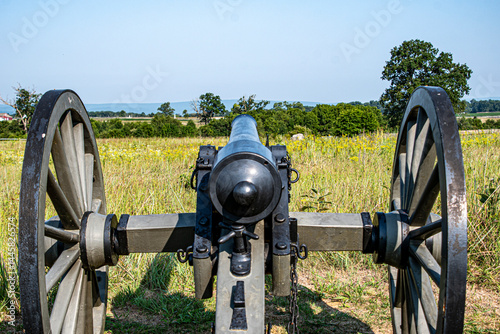 A vintage cannon stands still on a grassy knoll, pointing towards a sprawling battlefield. The sky shines blue, and trees frame the horizon, evoking a sense of history.