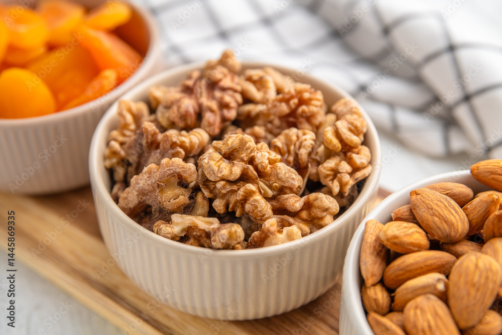 Three white ceramic bowls filled with almonds, walnuts, and dried apricots, arranged on a wooden cutting board, offer a healthy and delicious snack or ingredient for various culinary creations Three white ceramic bowls filled with almonds, walnuts, and dried apricots, arranged on a wooden cutting board, offer a healthy and delicious snack or ingredient for various culinary creations