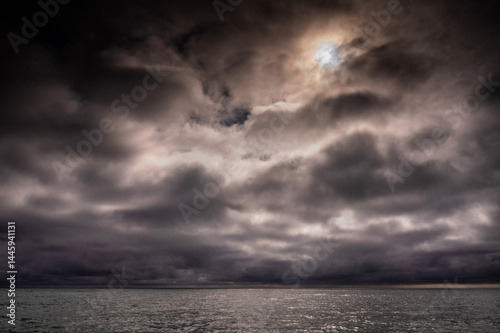 Obraz na płótnie Sun between dark storm clouds over ocean. Dramatic seascape. Clouds and rain flurry over sea. w salonie