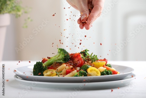 Fototapeta Naklejka Na Ścianę i Meble -  Woman sprinkling spices onto pasta with vegetables at white table indoors, closeup