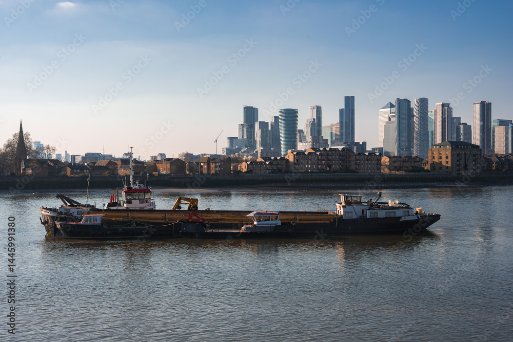 A barge floats on the River Thames with Canary Wharf's modern skyscrapers and a church spire visible, reflecting on calm water under a clear blue sky.