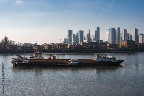 A barge floats on the River Thames with Canary Wharf's modern skyscrapers and a church spire visible, reflecting on calm water under a clear blue sky.