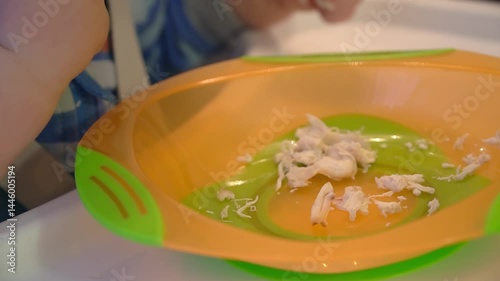 A one-year-old boy eats boiled chicken meat from a plastic plate with his hands. Teaching self-nutrition of a child. Hand motor skills development