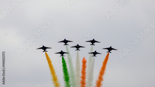 Aerial display of fighter jets leaving vibrant orange, white, and green smoke trails in the sky, representing the Indian flag. The planes fly in a precise V-formation against a pale sky.

