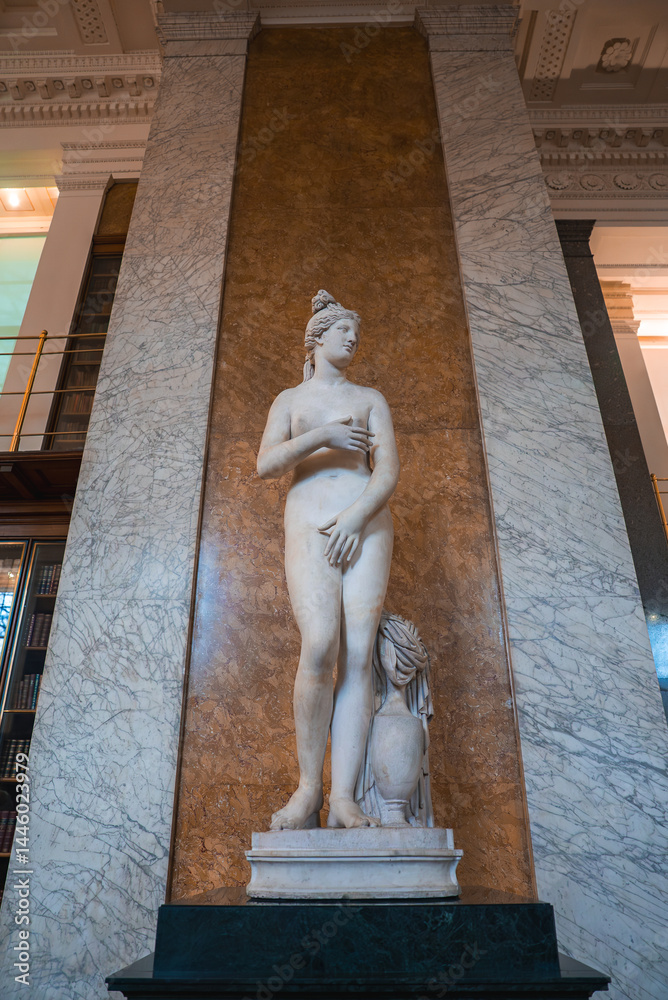 Classical marble statue of a nude female figure in contrapposto pose, set in a grand London interior with bookshelves and decorative elements.