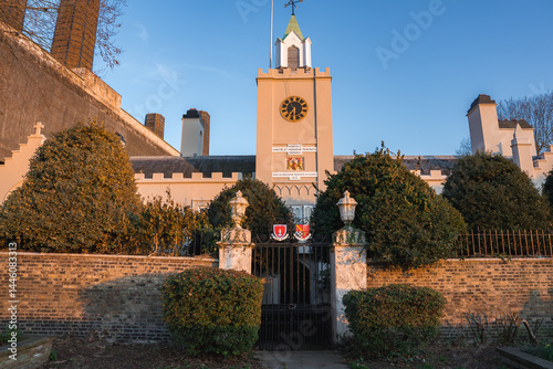 Obraz na płótnie The Royal Hospital Chelsea in London features a central clock tower, golden clock face, ornate iron gate, trimmed hedges, and a cream facade under blue skies. w salonie