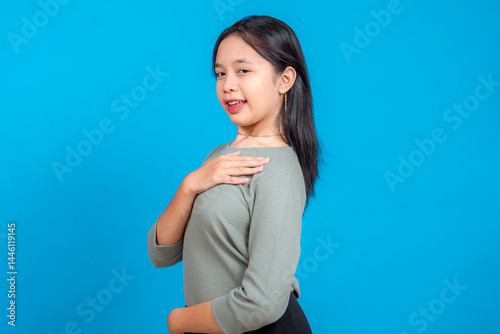 Fotografie Side view of a young Asian girl with a confident smirk and relaxed pose, one hand on her chest and the other on her waist, standing against a bright blue background