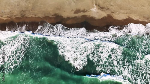 Aerial view capturing emerald green waves breaking over golden sand, white foam cascading along pristine coastal landscape with serene marine scenery