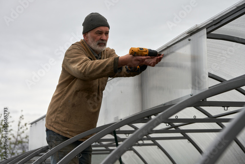 Covering the metal frame of the greenhouse with cellular polycarbonate. A man bolts self-tapping screws and screwdriver fastens polycarbonate to the frame of the upper windows.