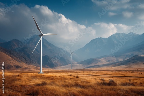 Wind turbines stand tall in a golden grassland with mountains shrouded in mist under a partly cloudy sky.