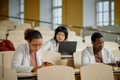Diverse group of med students sitting in atrium of lecture hall and taking notes during class in college