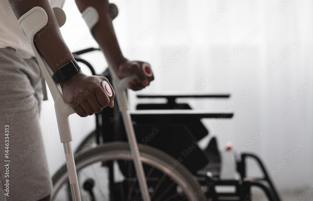 Obraz premium Physical activity and training to recover from injury and illness. Adult african american man disabled with crutches walks near wheelchair on large window background in interior, cropped, close up