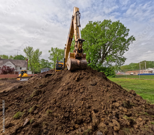 An industrial excavator, bulldozer or backhoe uses a hydraulic arm with a scoop to move dirt and grass in a field. City location with cloudy blue skies.