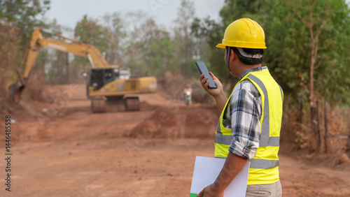 Obraz na płótnie Construction Supervision: An engineer stands at a construction site, communicating via radio, overseeing the project with attention to detail. Excavator and terrain present in scene. w salonie