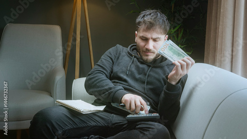 Stressed man calculating his remaining money and expenses at home, holding dolar banknotes and budgeting in notebook