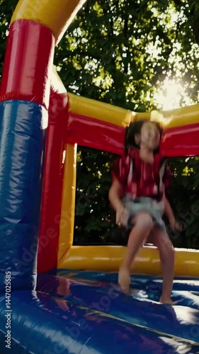 A joyful child jumps in a colorful bouncy castle. Captured from a low angle, the video exudes energy and fun against a backdrop of sunlit trees.